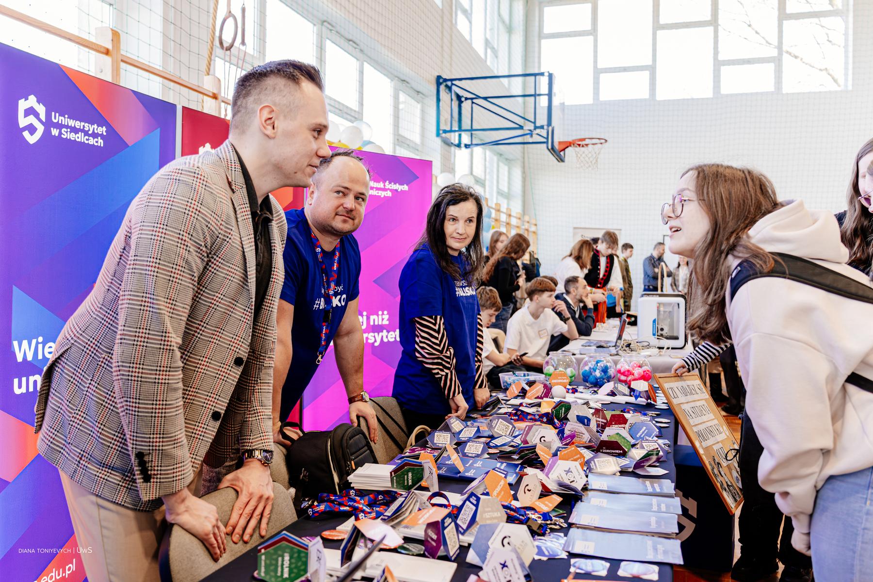 Pracownicy UwS promują uniwersytet na stoisku targów na sali gimnastycznej.